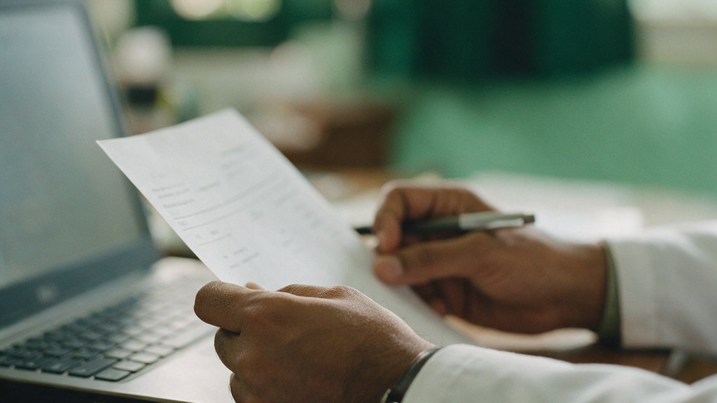 A doctor's hands holding a printed report beside an open laptop.